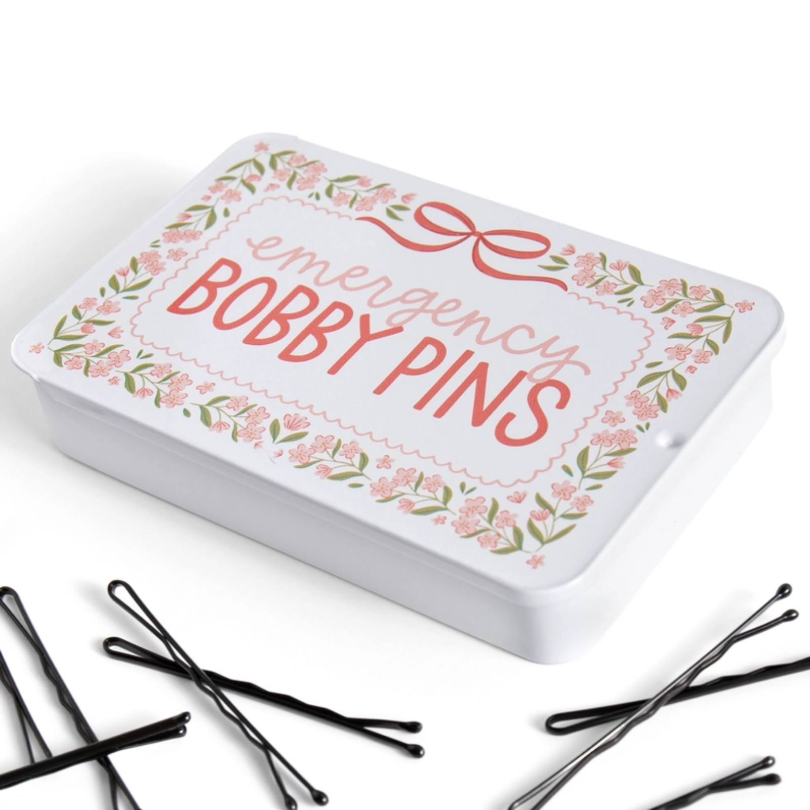 A white rectangular tin labeled 'emergency BOBBY PINS' with a pink bow and floral border, shown on a white background with several black bobby pins scattered in front.