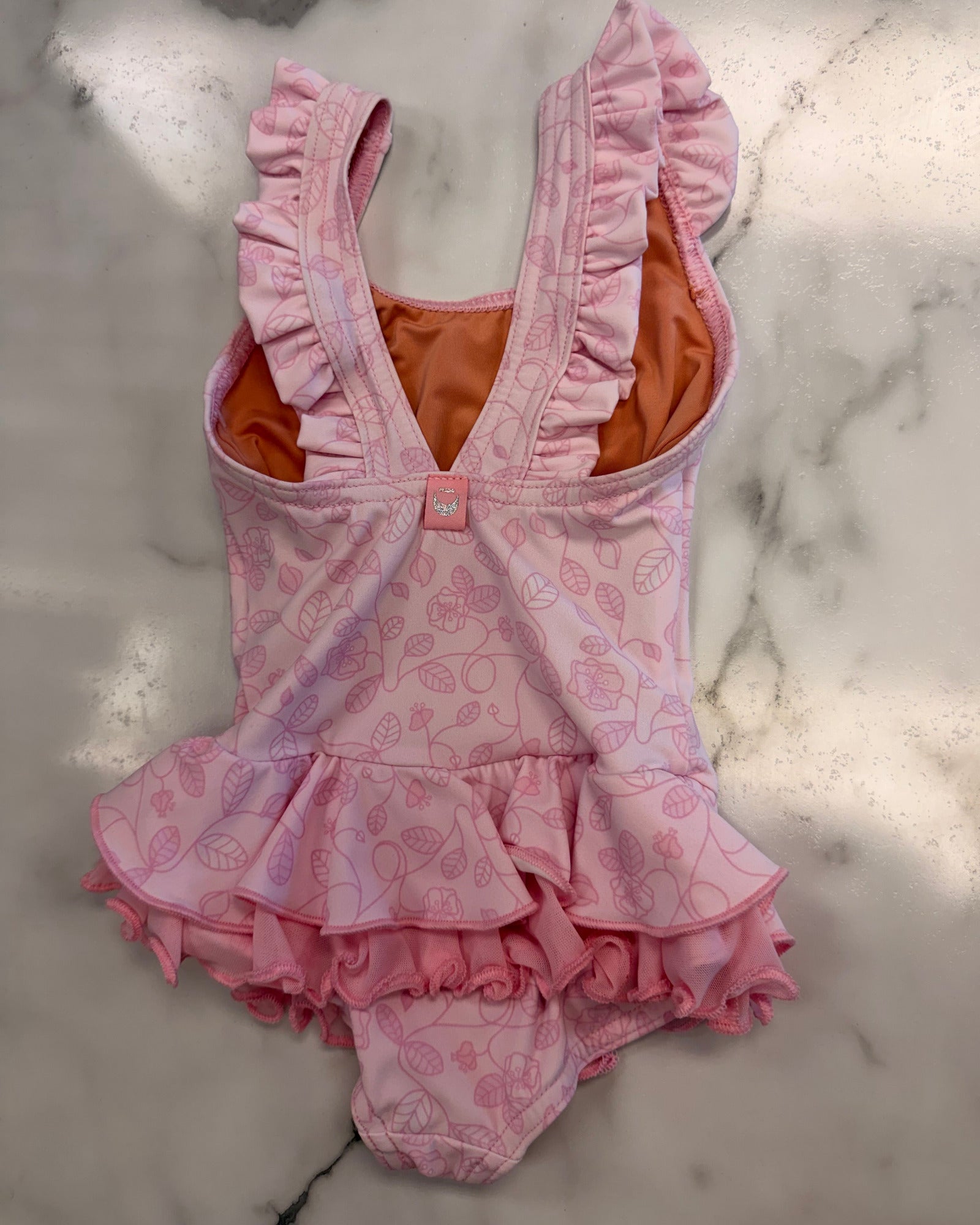 A pink baby girl's one-piece leotard with ruffled straps and a ruffled hem, featuring a light pink strawberry pattern and a bright orange inner lining, laid flat on a white marble surface.