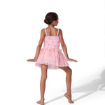 A young girl with her hair in a bun is shown from behind wearing a light pink tiered tutu dress with thin shoulder straps, posing on a solid white background.