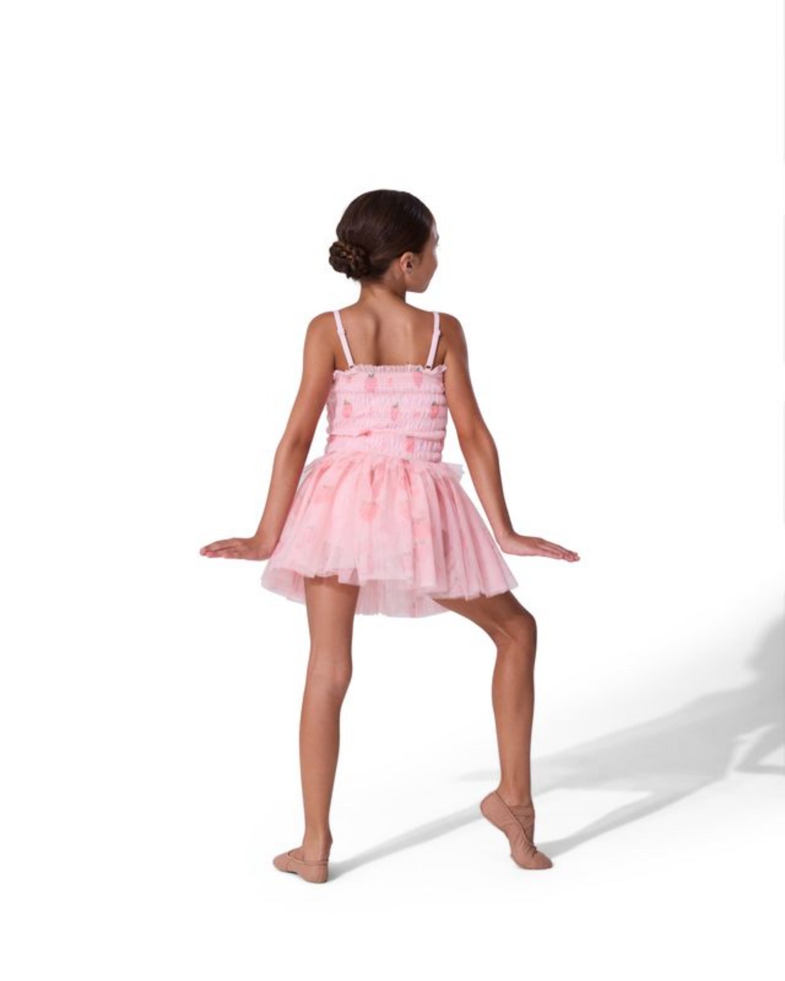 A young girl with her hair in a bun is shown from behind wearing a light pink tiered tutu dress with thin shoulder straps, posing on a solid white background.