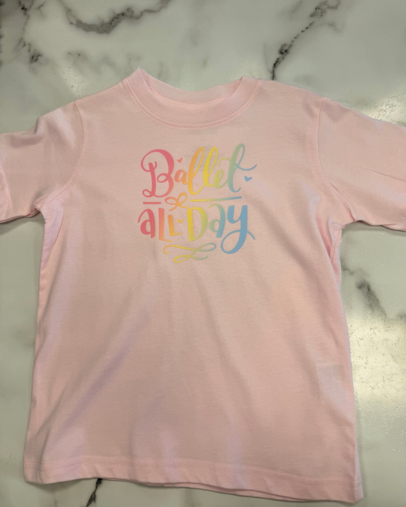 A light pink short-sleeve t-shirt with a colorful 'Ballin' all day' graphic printed on the chest, laid flat on a white and gray marble surface.
