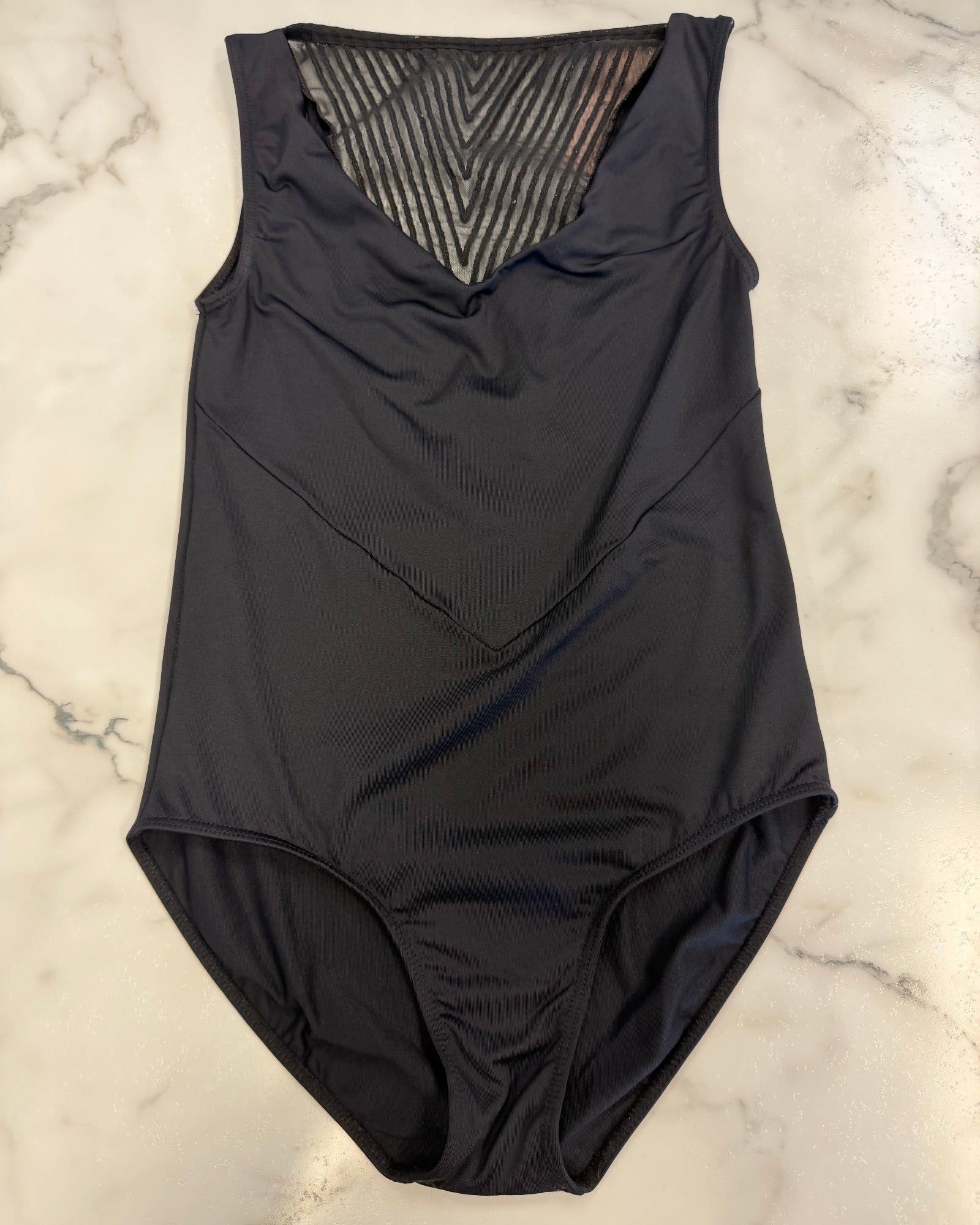 A black sleeveless leotard with a V-neckline and a textured mesh chest panel, laid flat on a white marble surface.