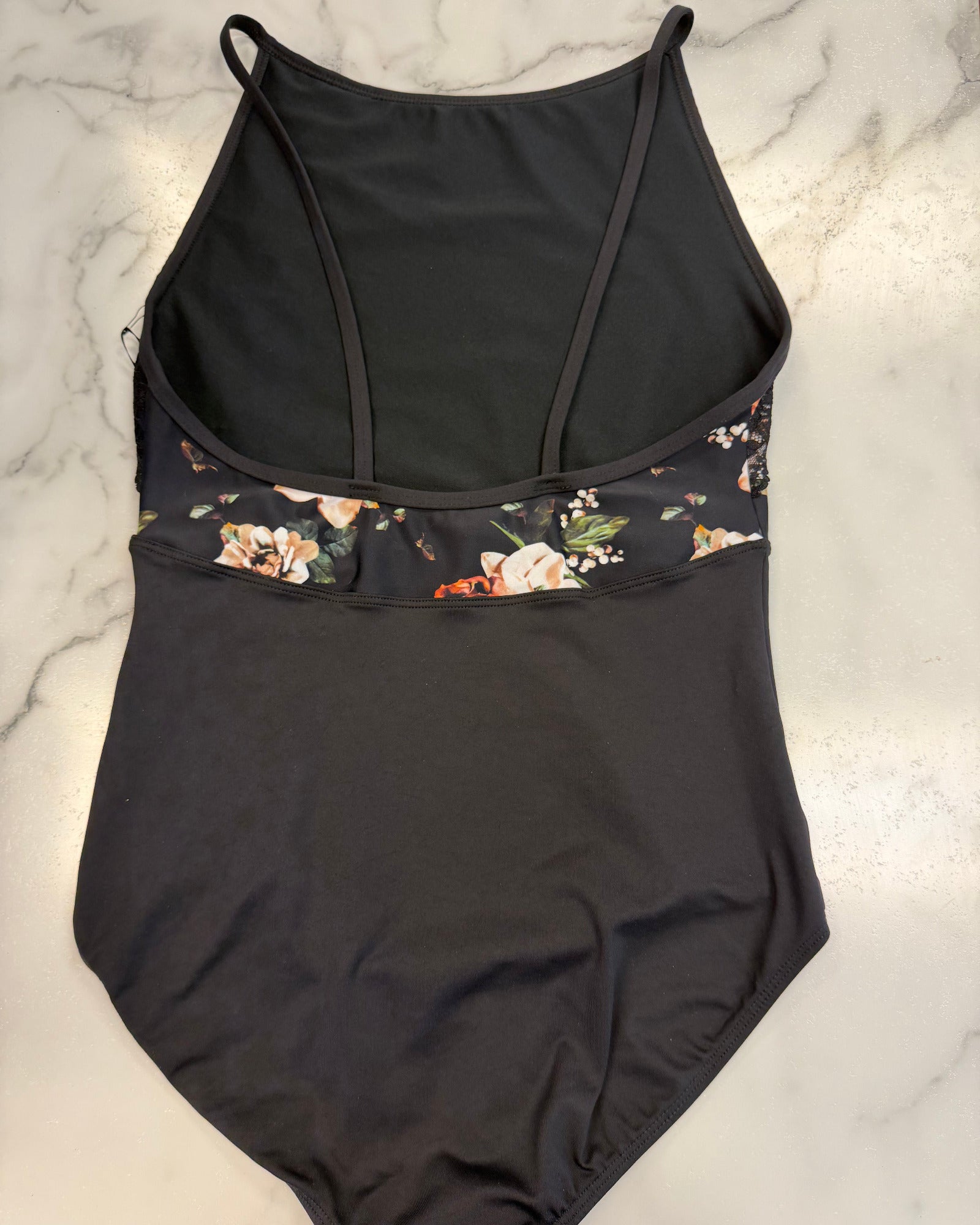 A black one-piece leotard with a floral patterned underbust band, laid flat on a white marble surface.