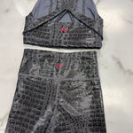A two-piece activewear set consisting of a sports bra and matching shorts, both featuring a dark gray and black crocodile-embossed pattern with a small pink logo on the front. The set is laid flat on a white marble background.