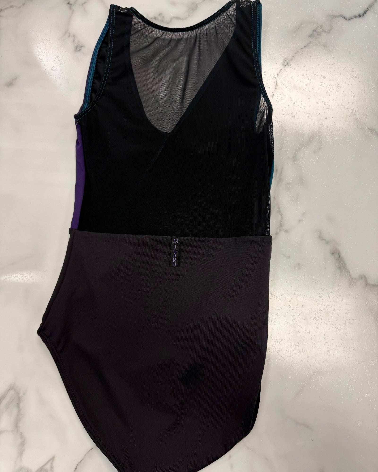 A black sleeveless leotard laid flat on a white marble surface, featuring a sheer mesh V-neck back panel, purple side panels, and teal trim.
