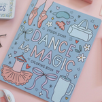 A flat-lay photograph of the 'Dance is Magic Coloring Book' by Pippi Post, featuring a light blue cover decorated with pink and white illustrations of ballet shoes, a tutu, and bows. The book is styled on a pink background with various cosmetic and accessory props.