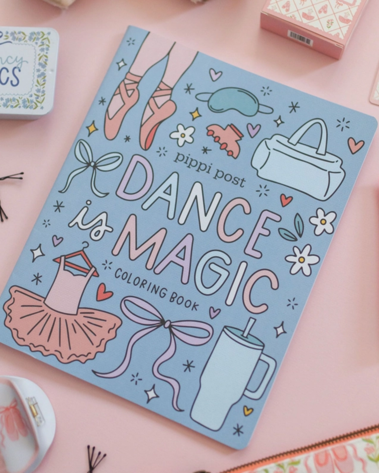 A flat-lay photograph of the 'Dance is Magic Coloring Book' by Pippi Post, featuring a light blue cover decorated with pink and white illustrations of ballet shoes, a tutu, and bows. The book is styled on a pink background with various cosmetic and accessory props.
