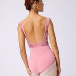 A woman modeling a pink dance leotard from the back, showing a lace-trimmed open back and high waist, paired with nude tights against a light gray studio background.
