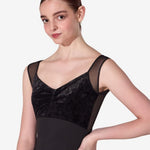 A female model wearing a black dance leotard with a velvet bodice, sheer mesh shoulder panels, and a solid black skirt, posed against a light gray studio background.