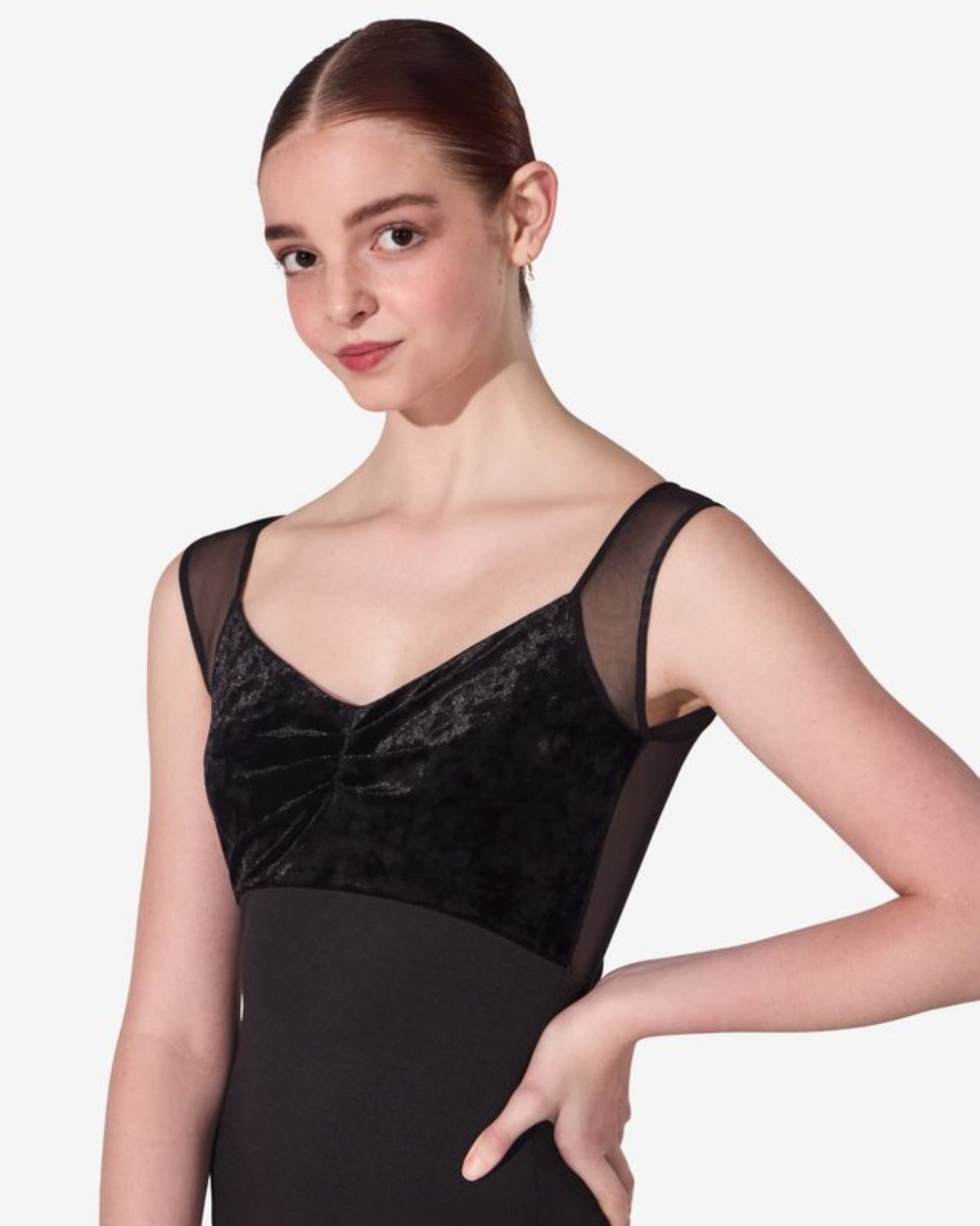 A female model wearing a black dance leotard with a velvet bodice, sheer mesh shoulder panels, and a solid black skirt, posed against a light gray studio background.