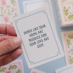 A hand holds a square white card with a light blue border and the phrase 'DANCE LIKE YOUR ARMS ARE NOODLES AND YOUR LEGS ARE STIFF.' in dark blue text.