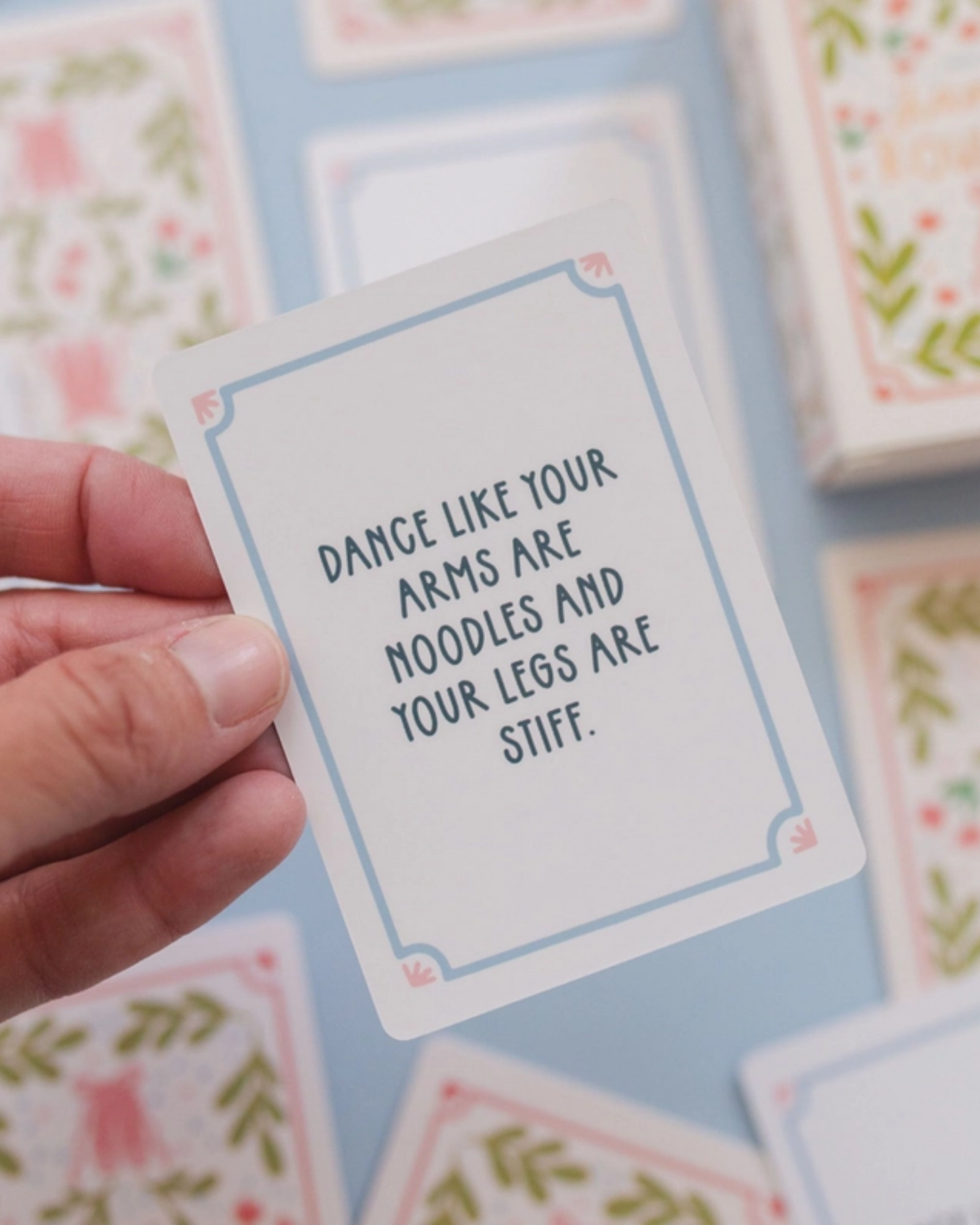 A hand holds a square white card with a light blue border and the phrase 'DANCE LIKE YOUR ARMS ARE NOODLES AND YOUR LEGS ARE STIFF.' in dark blue text.