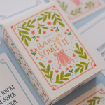 A decorative product box labeled 'dancer ROULETTE' sits on a light blue surface with matching printed cards partially visible around it. The box has a white base with green leafy borders, pink floral accents, and a pink dancer silhouette in the center.