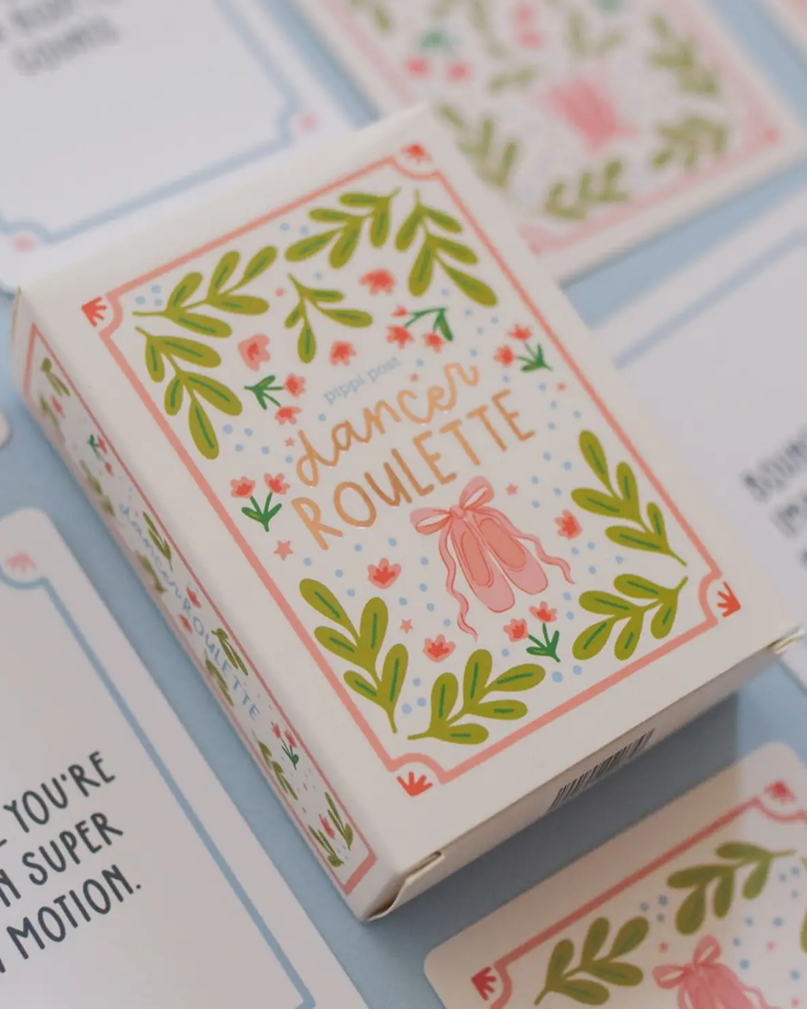 A decorative product box labeled 'dancer ROULETTE' sits on a light blue surface with matching printed cards partially visible around it. The box has a white base with green leafy borders, pink floral accents, and a pink dancer silhouette in the center.