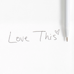 A close-up of a white pen on a white background next to handwritten text reading 'Love This' with a small heart drawn in the letter 'i'.