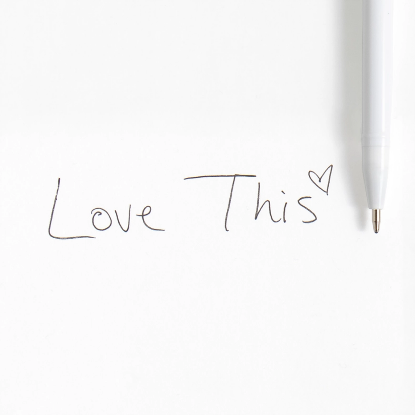 A close-up of a white pen on a white background next to handwritten text reading 'Love This' with a small heart drawn in the letter 'i'.