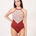 A woman modeling a leotard with a rust red color, featuring a floral lace pattern on the bust and halter-style straps, set against a plain light background.