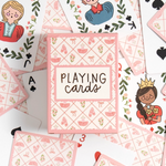 A styled flat-lay of a pink and white playing card deck box surrounded by illustrated playing cards on a white background. The box reads 'PLAYING cards' and features a pink diamond pattern with small shoe motifs.