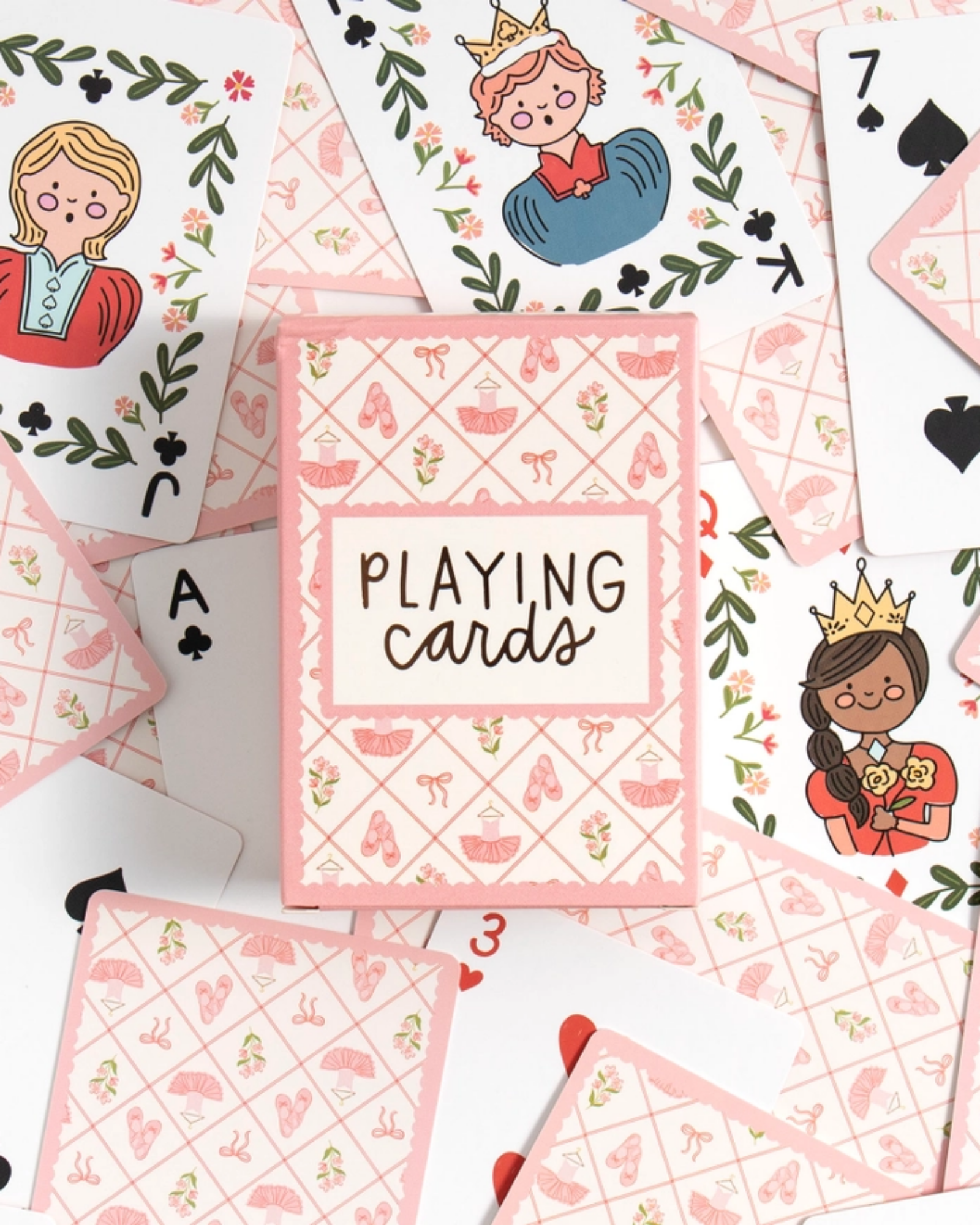 A styled flat-lay of a pink and white playing card deck box surrounded by illustrated playing cards on a white background. The box reads 'PLAYING cards' and features a pink diamond pattern with small shoe motifs.