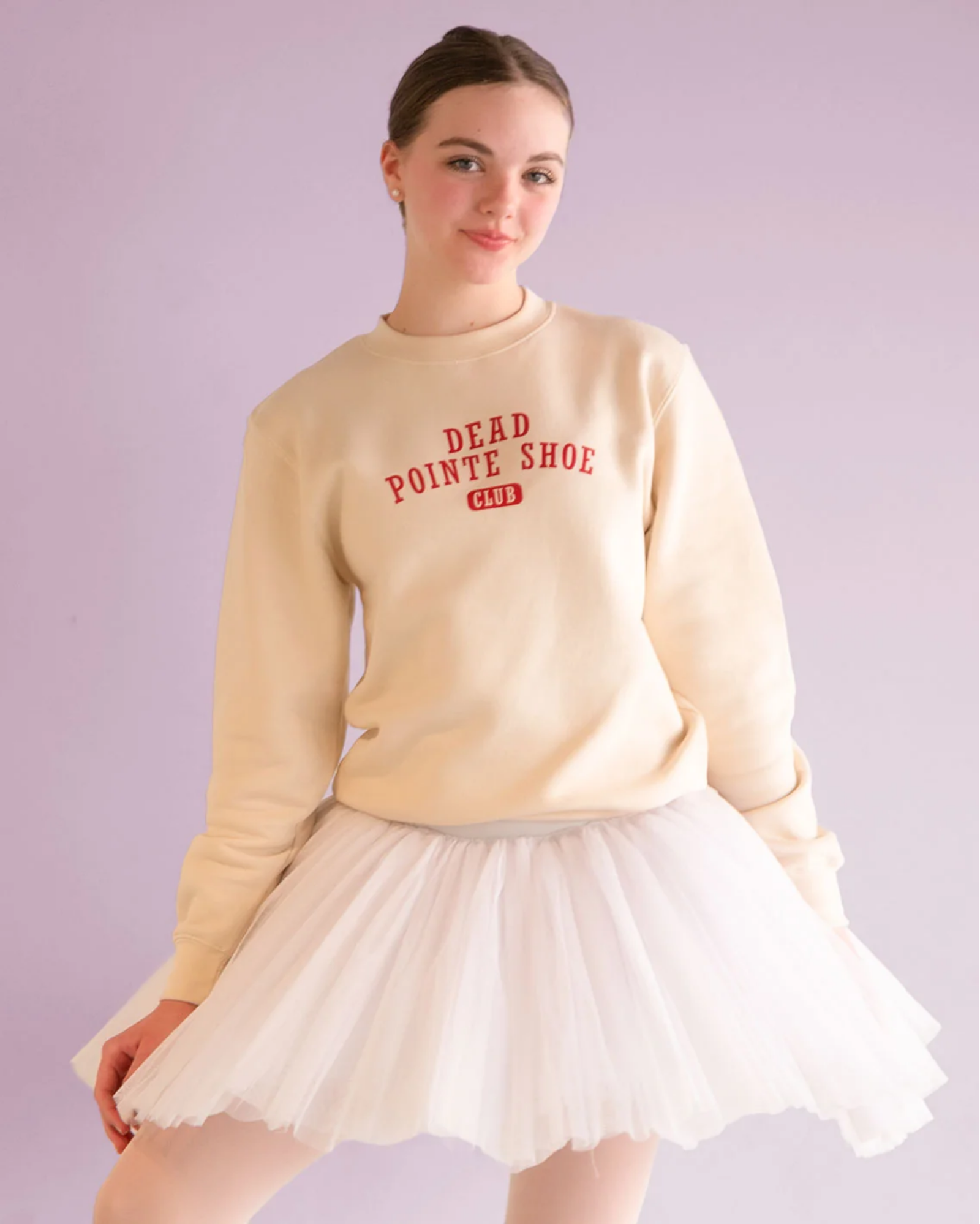 Woman wearing a beige sweatshirt with text and a white skirt on a light purple background