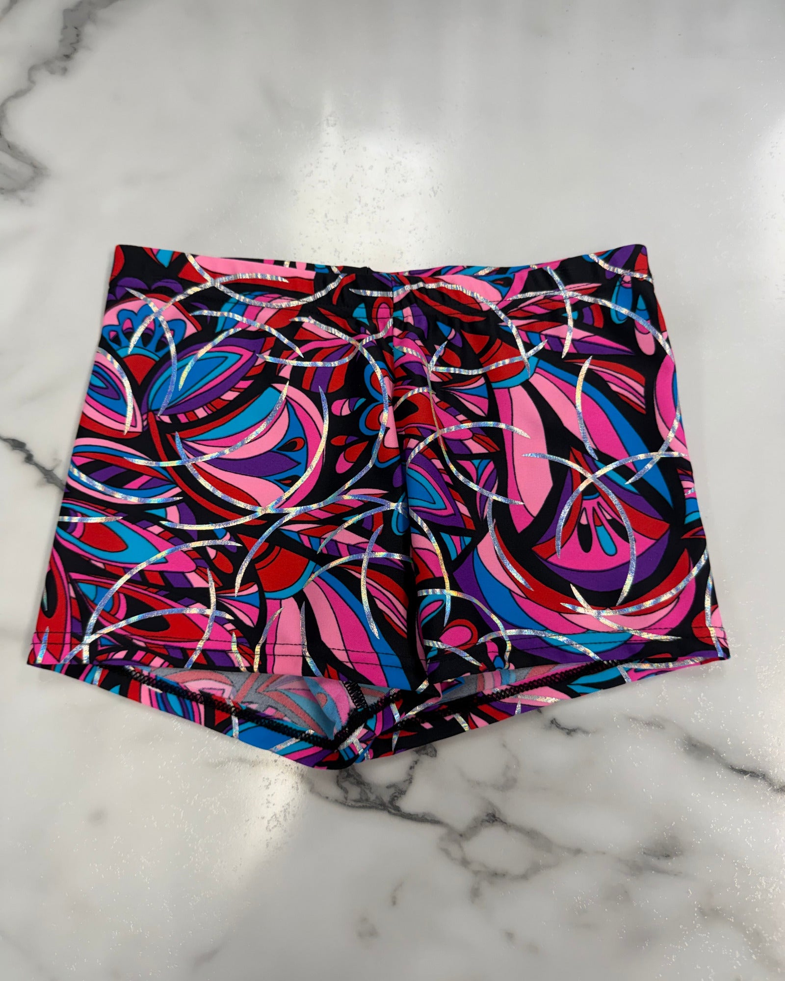 A pair of dance shorts featuring a vibrant, abstract psychedelic swirl pattern in pink, blue, red, and black, laid flat on a white marble surface.