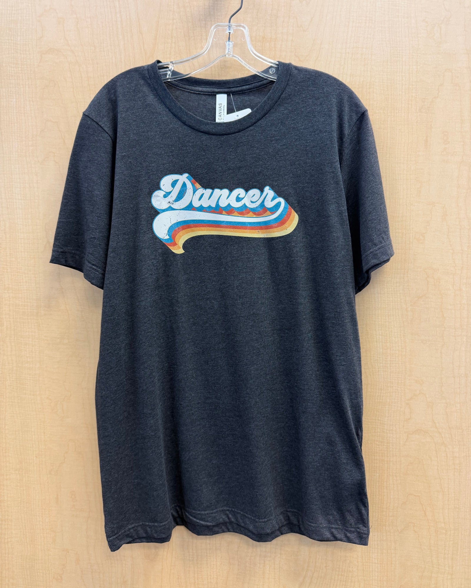 A dark charcoal gray heather t-shirt hanging on a clear plastic hanger against a light wood-paneled wall. The shirt features a retro-style graphic on the chest with the word 'Dancer' in white script over a wavy blue, orange, and yellow stripe design.
