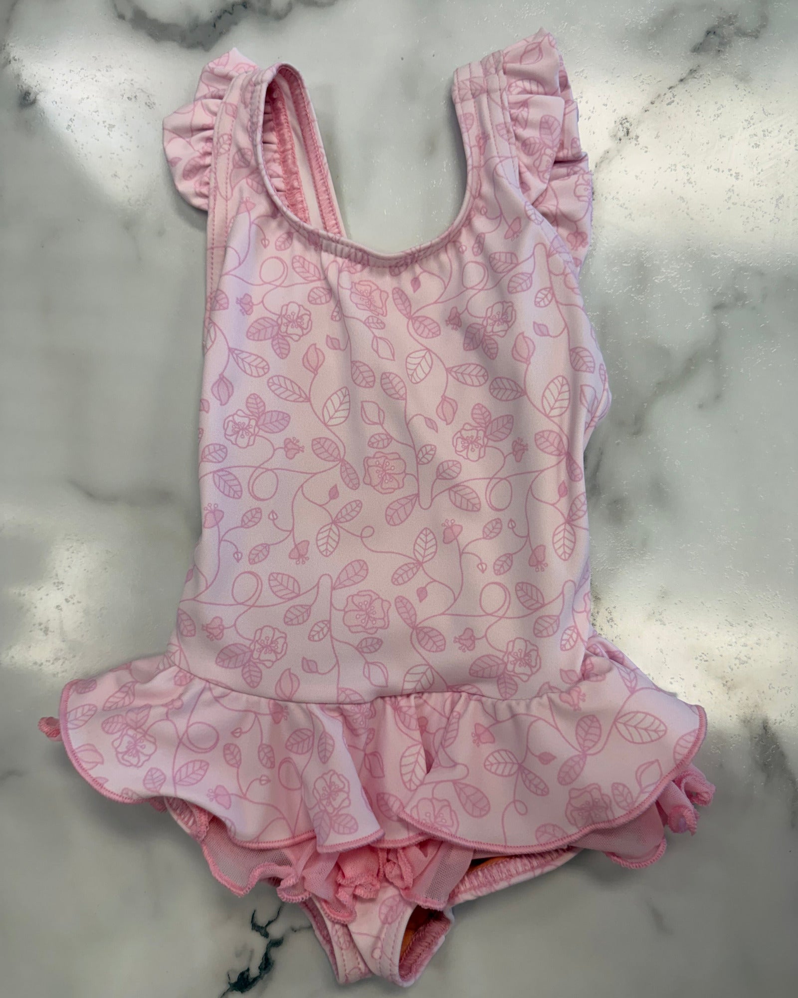 A pink one-piece baby leotard with a delicate pink floral and leaf pattern, featuring ruffled shoulder straps and a ruffled hem, laid flat on a white marble surface.
