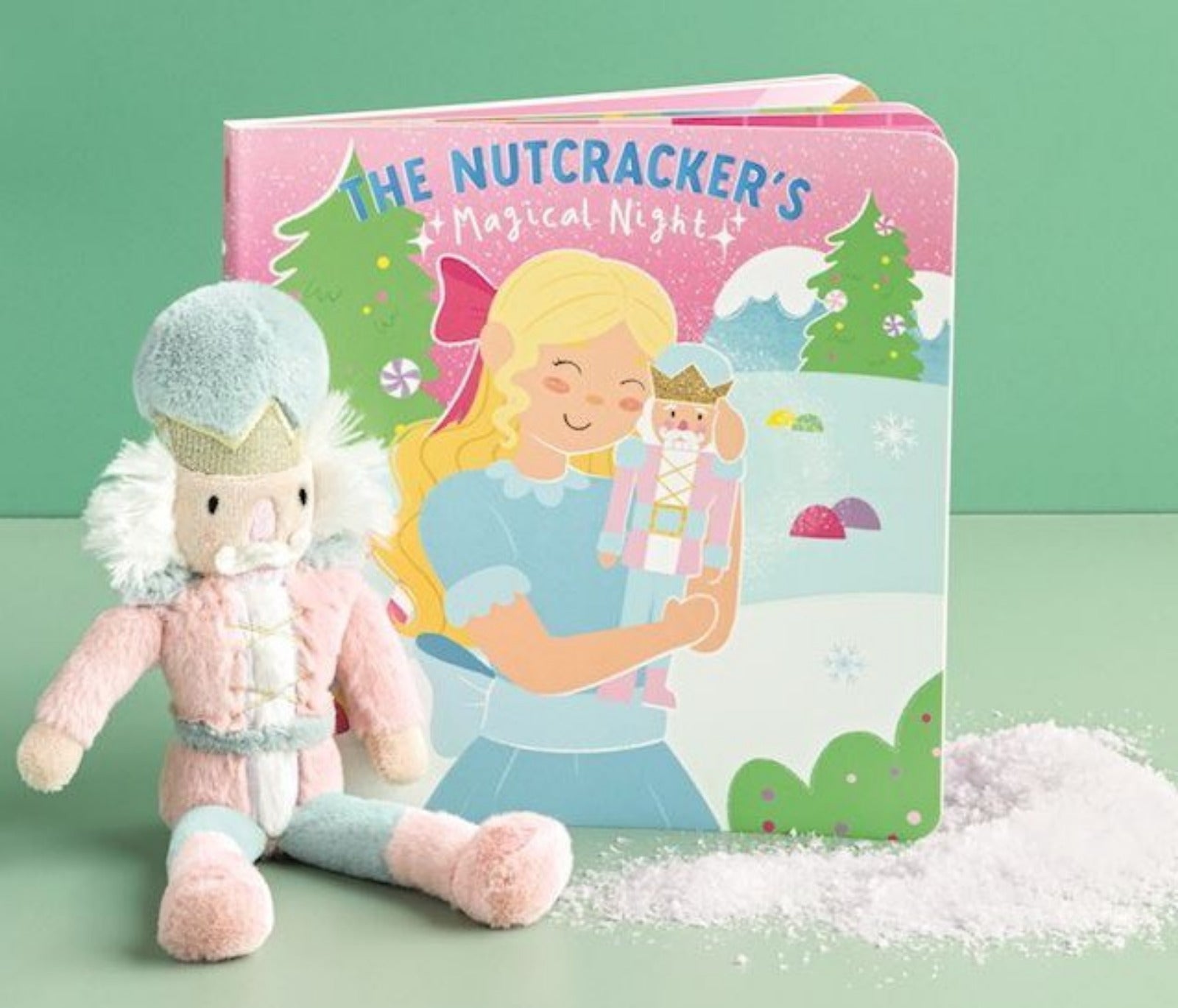 A children's book titled "The Nutcracker's Magical Night" stands upright next to a plush nutcracker doll, with a small pile of white faux snow on a mint green surface and background.