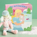 A children's book titled "The Nutcracker's Magical Night" stands upright next to a plush nutcracker doll, with a small pile of white faux snow on a mint green surface and background.