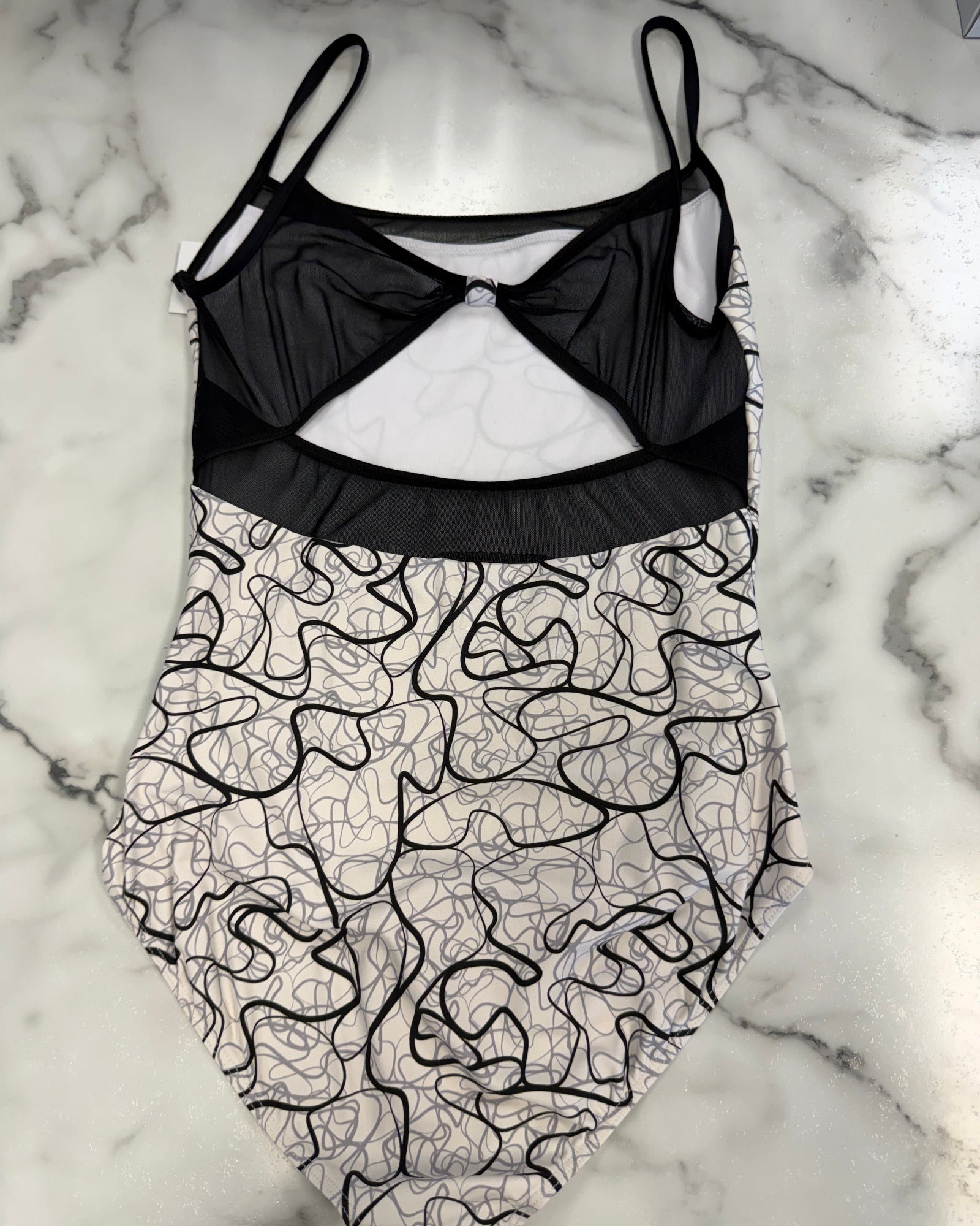 A leotard with a black mesh bustier-style top and a white lower body covered in a black abstract line pattern, laid flat on a white and gray marble surface.
