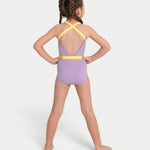 A young girl with braided hair is shown from the back wearing a lavender purple leotard with yellow straps and a yellow waistband, standing against a light gray background.