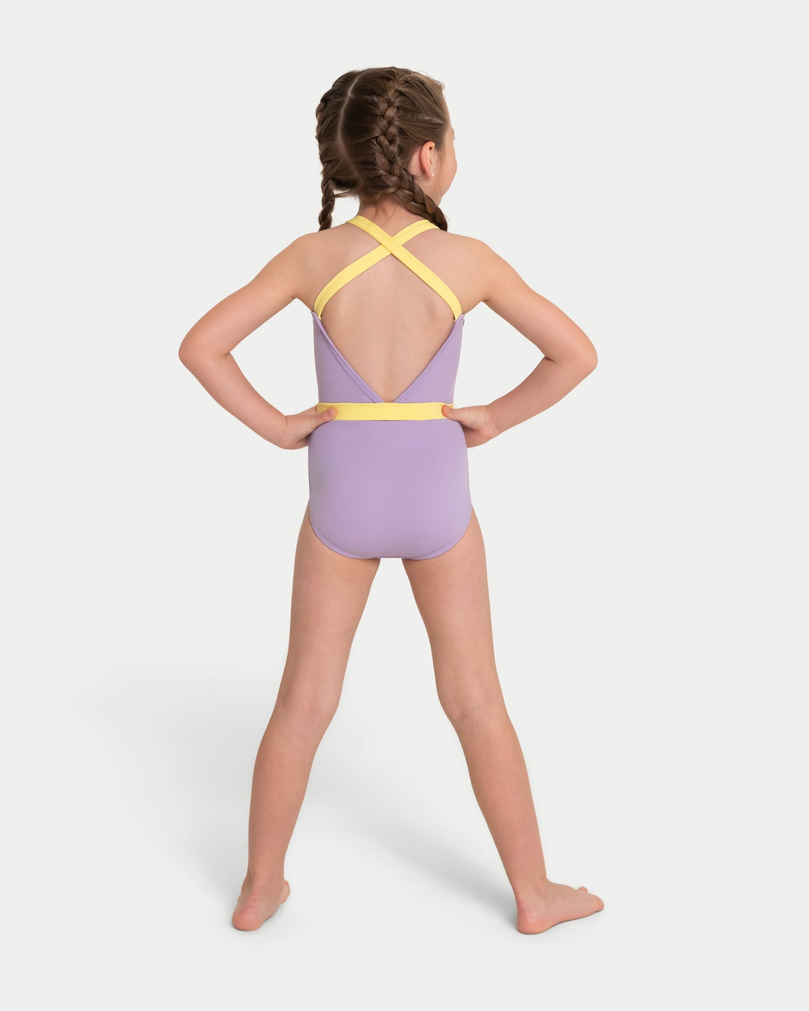 A young girl with braided hair is shown from the back wearing a lavender purple leotard with yellow straps and a yellow waistband, standing against a light gray background.