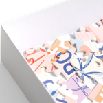 A close-up of a white box containing colorful jigsaw puzzle pieces with pastel graphics and lettering on a clean white background.