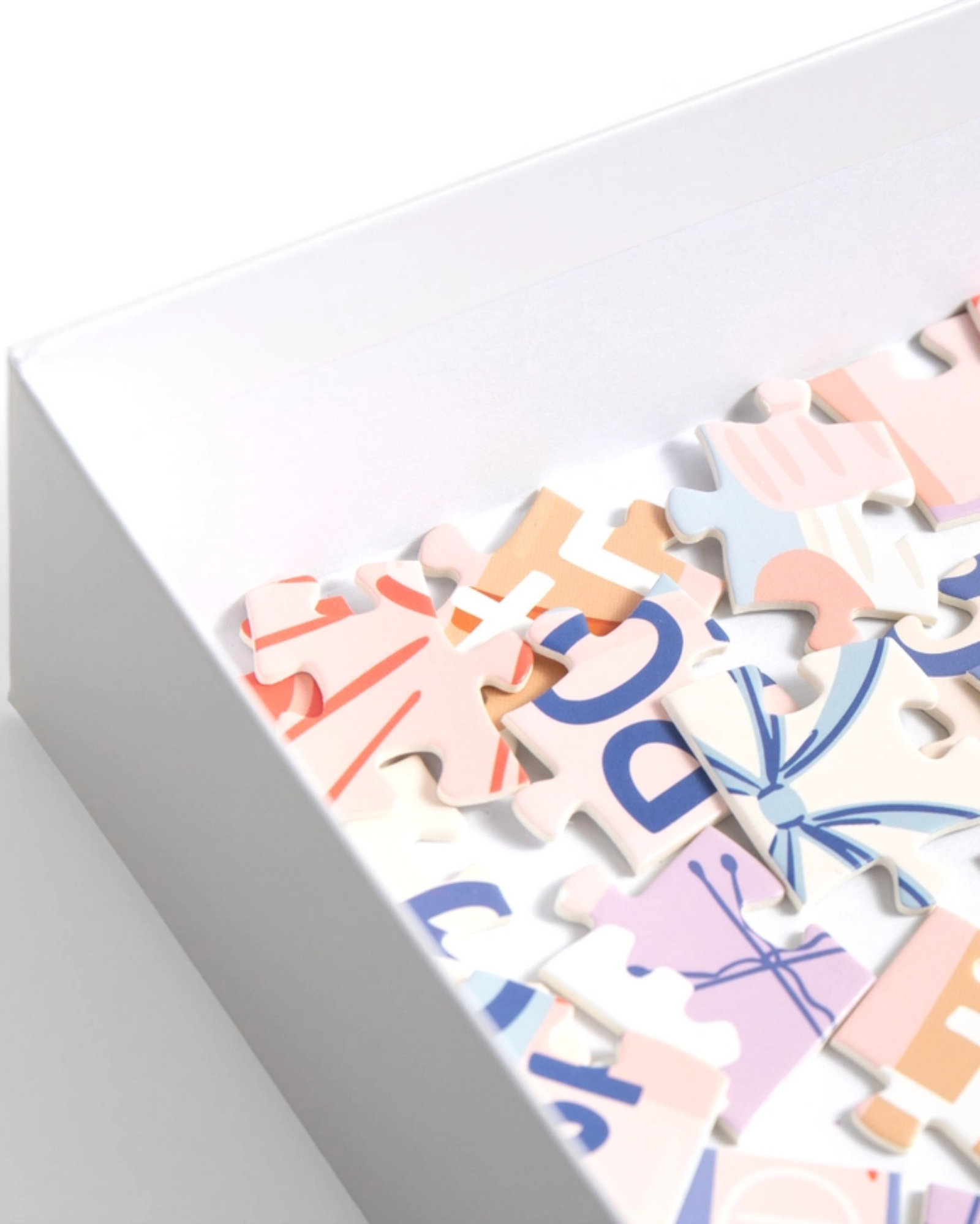 A close-up of a white box containing colorful jigsaw puzzle pieces with pastel graphics and lettering on a clean white background.