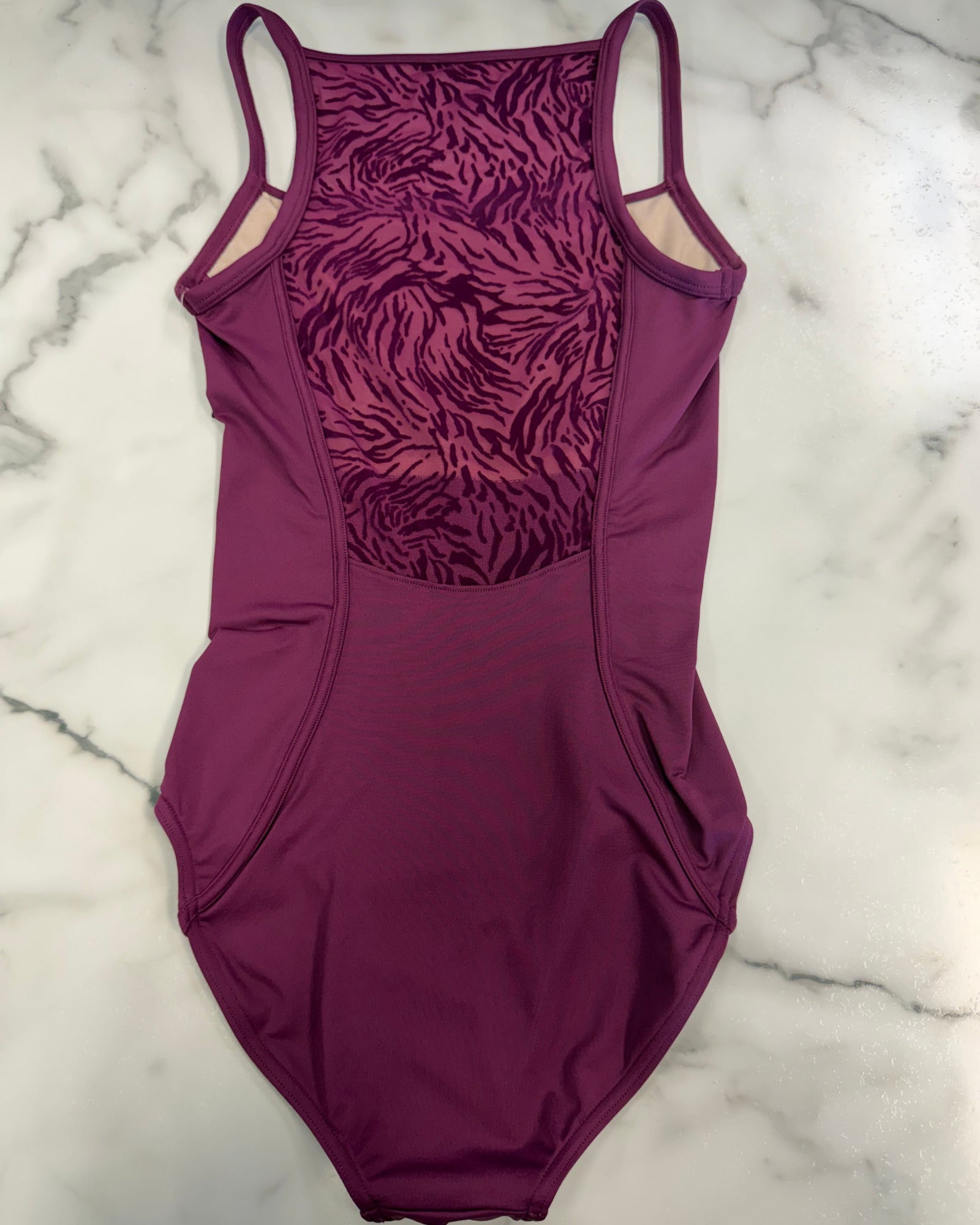 A women's leotard in a deep magenta-purple color, featuring a textured patterned panel on the back and thin shoulder straps, laid flat on a white marble surface.
