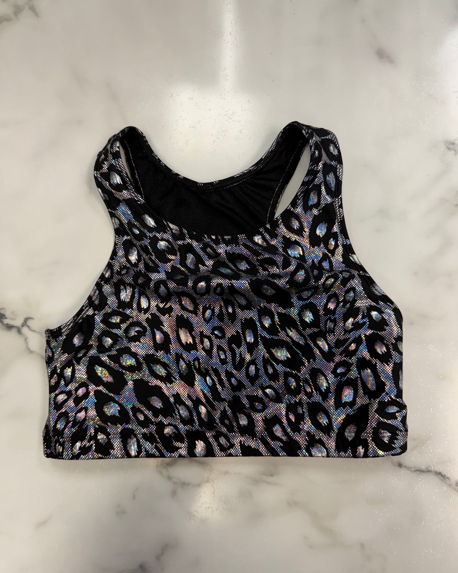 A sleeveless crop top with an iridescent leopard print pattern on a black background, laid flat on a white marble surface.