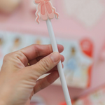 Hand holding a white pen with a pink ballerina clip against a blurred background