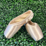 A pair of satin pointe shoes in a light champagne beige color, displayed on a textured green leaf wall background.