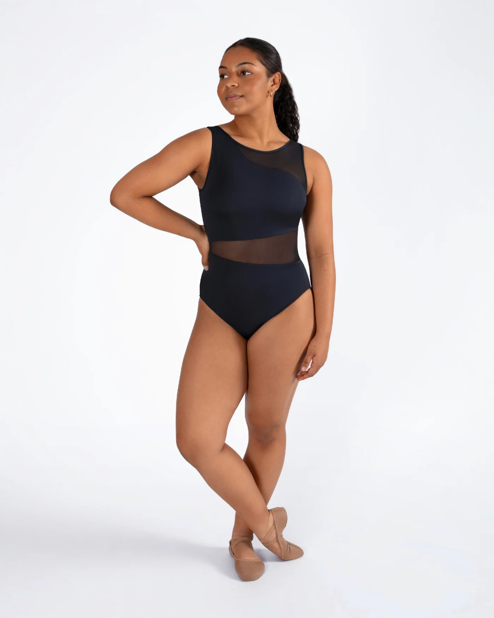 A woman modeling a sleeveless black dance leotard with sheer mesh panels, standing in ballet slippers against a plain white studio background.