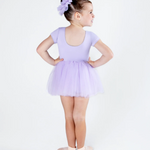 A young girl wearing a light purple short-sleeve leotard and matching tulle tutu, paired with a matching purple hair bow and pink ballet slippers, standing in profile against a white studio background.