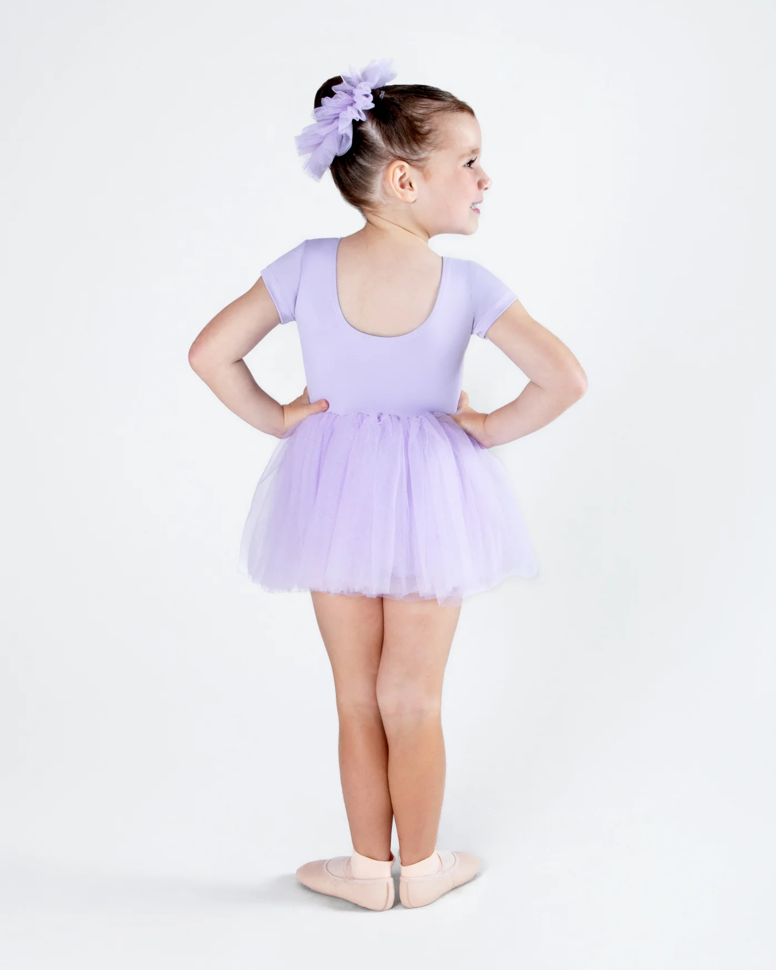 A young girl wearing a light purple short-sleeve leotard and matching tulle tutu, paired with a matching purple hair bow and pink ballet slippers, standing in profile against a white studio background.