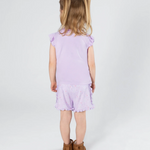 A young girl with blonde pigtails is shown from behind wearing a light purple ruffle-sleeve top and matching patterned shorts, paired with brown boots, against a plain light gray studio background.