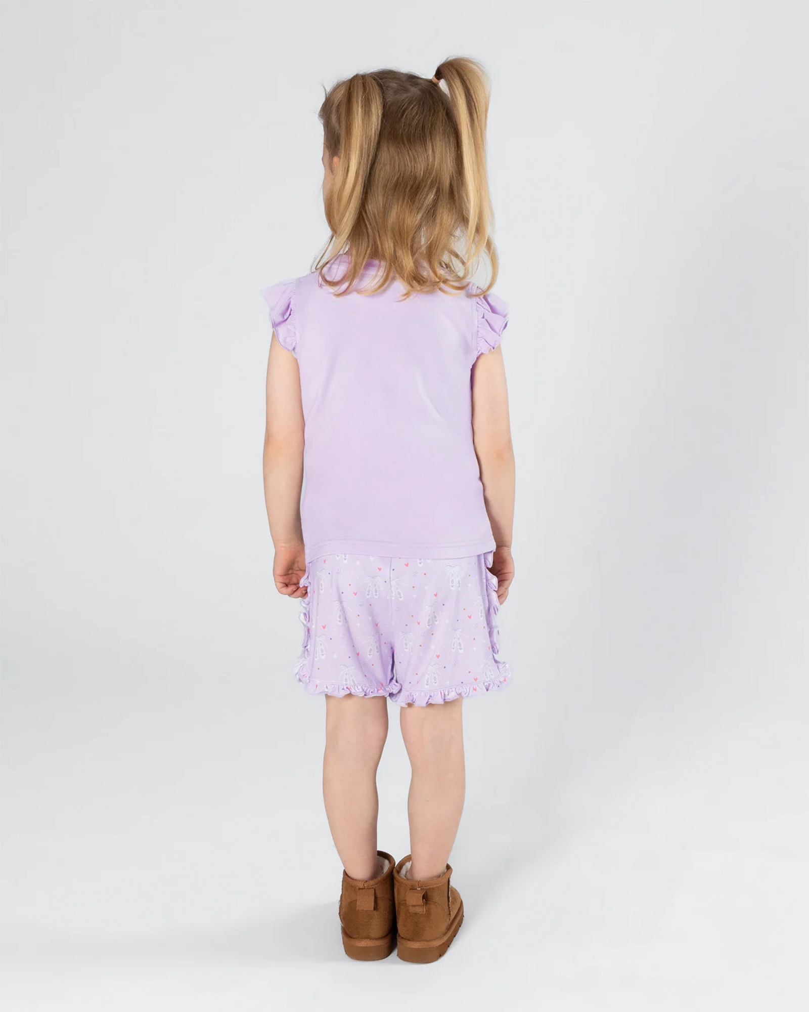 A young girl with blonde pigtails is shown from behind wearing a light purple ruffle-sleeve top and matching patterned shorts, paired with brown boots, against a plain light gray studio background.