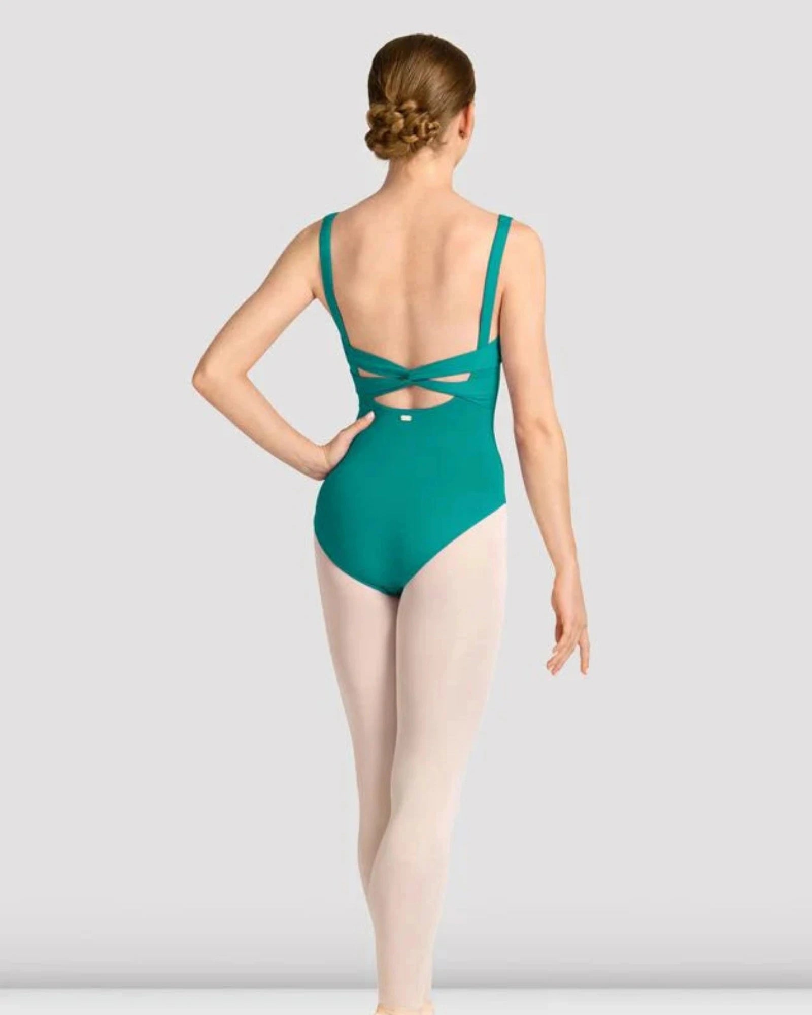 A female dancer shown from the back wearing a teal green leotard with a crossover back design, paired with light pink tights and pointe shoes, against a light gray studio background.