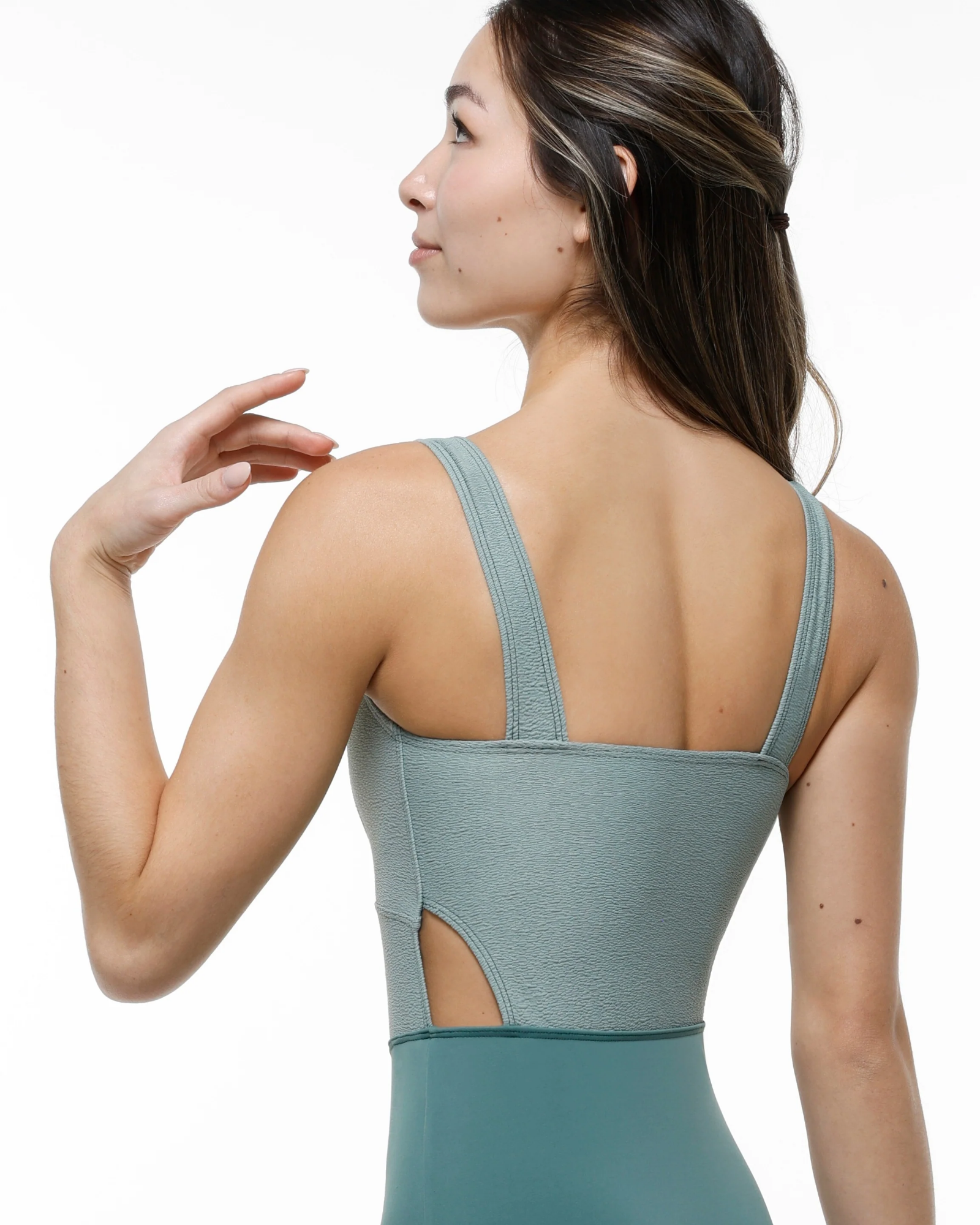 A woman shown from the back wearing a sage green leotard with wide shoulder straps and a side cutout, photographed against a clean white background.