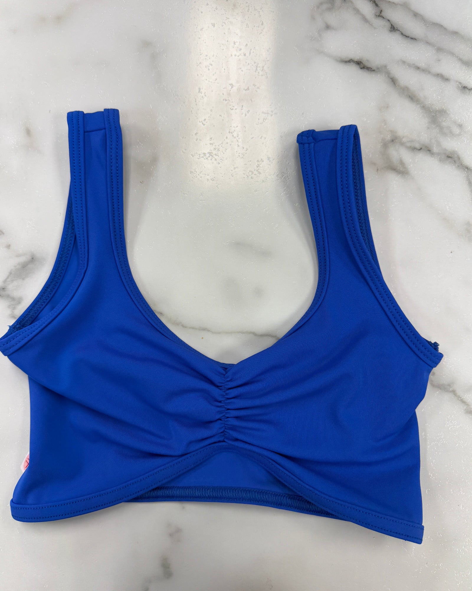 A bright royal blue dance bra with a ruched front detail, laid flat on a white marble surface.