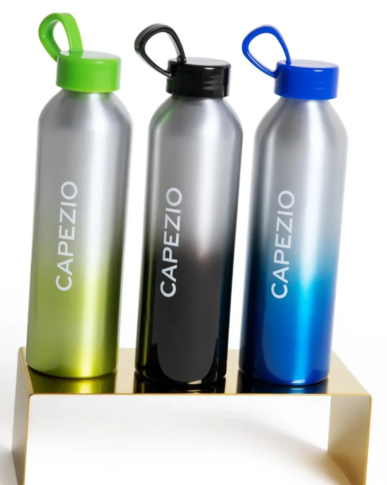 Three aluminum water bottles with gradient finishes in green, black, and blue, each with a matching colored loop cap and the brand name 'CAPEZIO' printed vertically, displayed on a gold-colored metal stand against a white background.