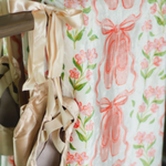 A close-up of a fleece blanket with a pink ballet ribbon and green floral print, hanging from a wooden rod with several satin ballet slippers tied to it.