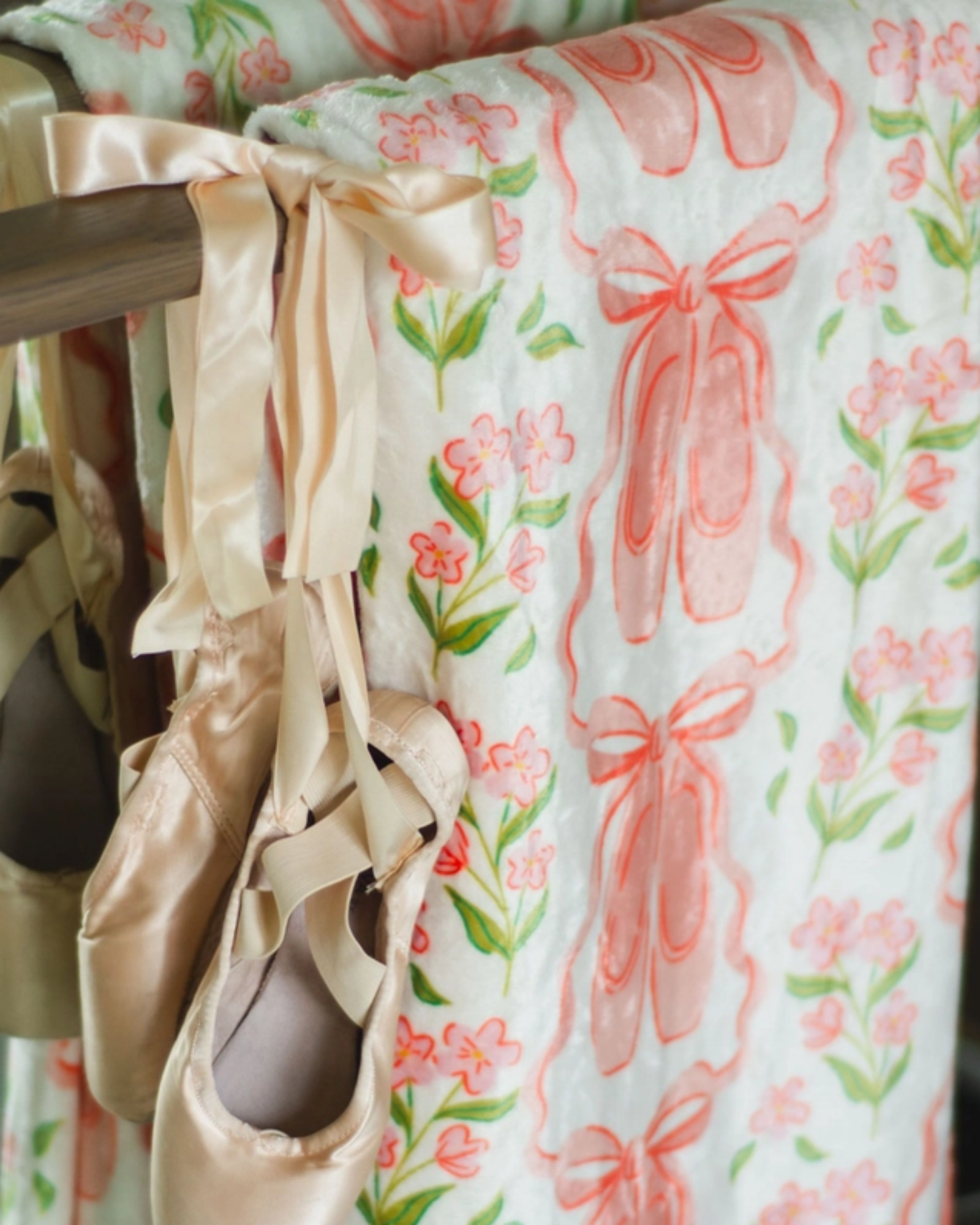 A close-up of a fleece blanket with a pink ballet ribbon and green floral print, hanging from a wooden rod with several satin ballet slippers tied to it.