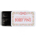 A rectangular metal tin of black bobby pins with a white lid labeled 'emergency BOBBY PINS' in coral pink text, surrounded by a floral border. The tin is shown open on a plain white background.